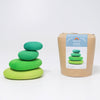 A set of four green and teal wooden stacking stones, crafted as non-toxic wooden toys, is arranged in a balanced tower on the left. On the right, a brown cylindrical package displays an image of the same stacked stones, labeled "Moss Pebbles Stacker." The background is plain white.