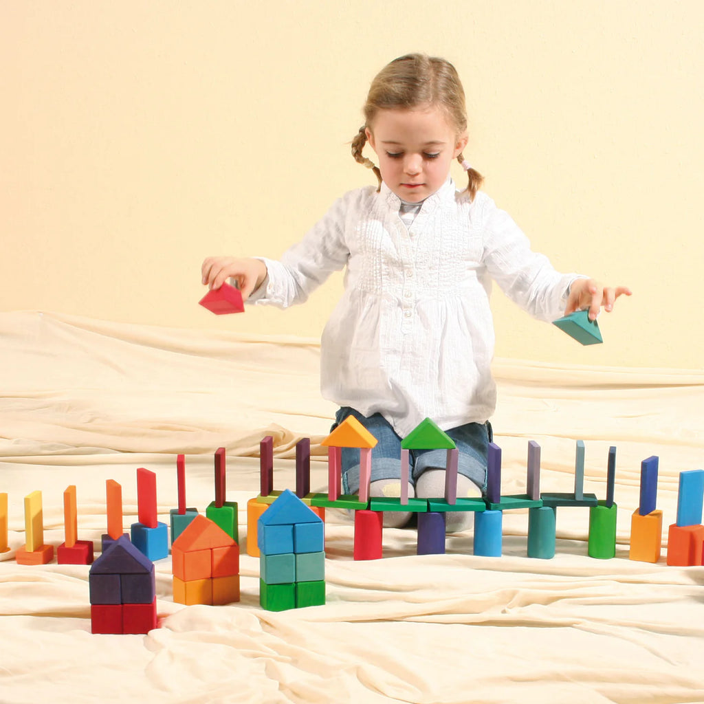 A young girl sits on a soft surface, arranging Grimm’s Shapes and Colors Set to build colorful structures and patterns, helping develop her fine motor skills.