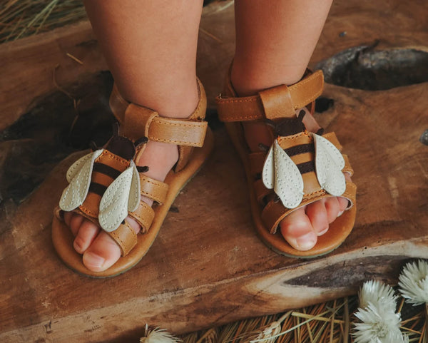 A close-up of a child's feet in Donsje Baby Bee Shoes with white leaf designs, standing on a rustic wood surface near pine needles.
