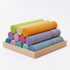Grimm's Large Building Rollers - Pastel (coming soon) features a wooden tray with pastel green, orange, purple, pink, and blue cylindrical blocks. Stacked in a pyramid, this engaging game boosts motor skills and stands out on a white background.