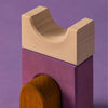 A close-up of stacked wooden toys from Grimm's Panorama Building Set on a purple surface, showing a natural wood-colored U-shaped block on a rectangular purple block, with a curved brown piece partially visible underneath.