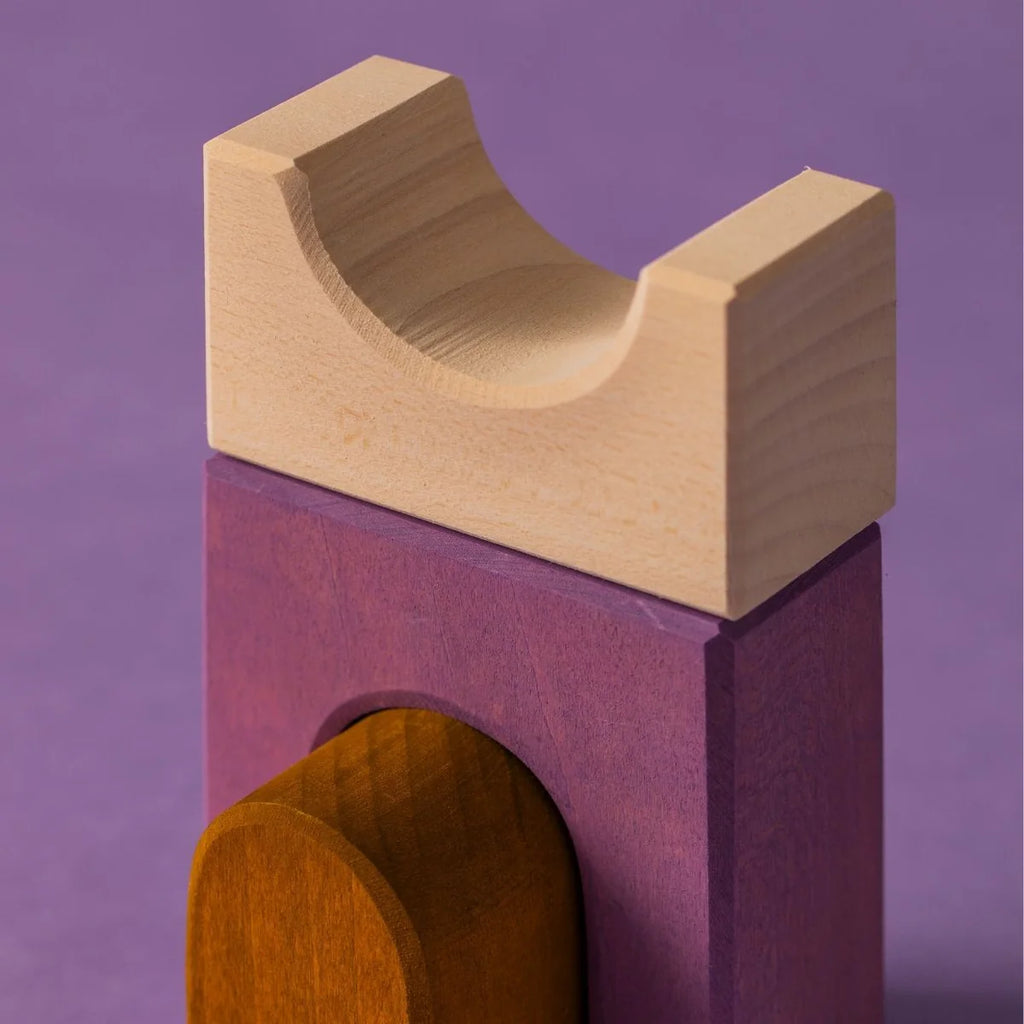 A close-up of stacked wooden toys from Grimm's Panorama Building Set on a purple surface, showing a natural wood-colored U-shaped block on a rectangular purple block, with a curved brown piece partially visible underneath.