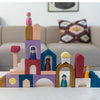 The Grimm's Panorama Building Set, featuring colorful wooden blocks and peg dolls, creates a whimsical cityscape on a table, while a beige couch with patterned pillows sits in the background.