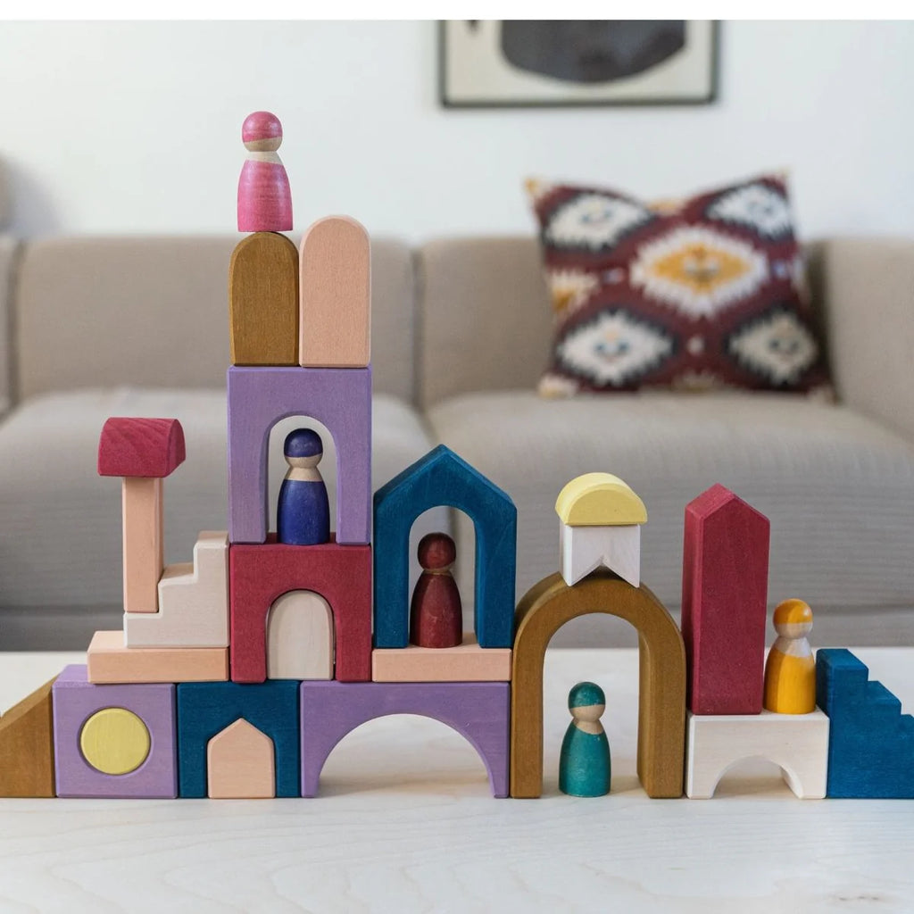 The Grimm's Panorama Building Set, featuring colorful wooden blocks and peg dolls, creates a whimsical cityscape on a table, while a beige couch with patterned pillows sits in the background.