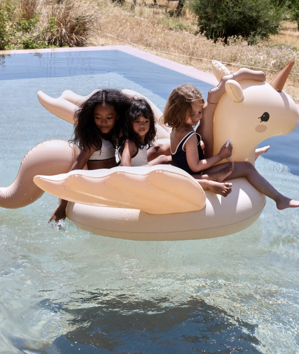 Three smiling children of diverse backgrounds enjoy a sunny day in a pool, riding together on a large Inflatable Unicorn Float.
