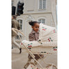 A young girl in a plaid dress stands by a white vintage style doll pram with cherry designs, on a city street with European architecture in the background.