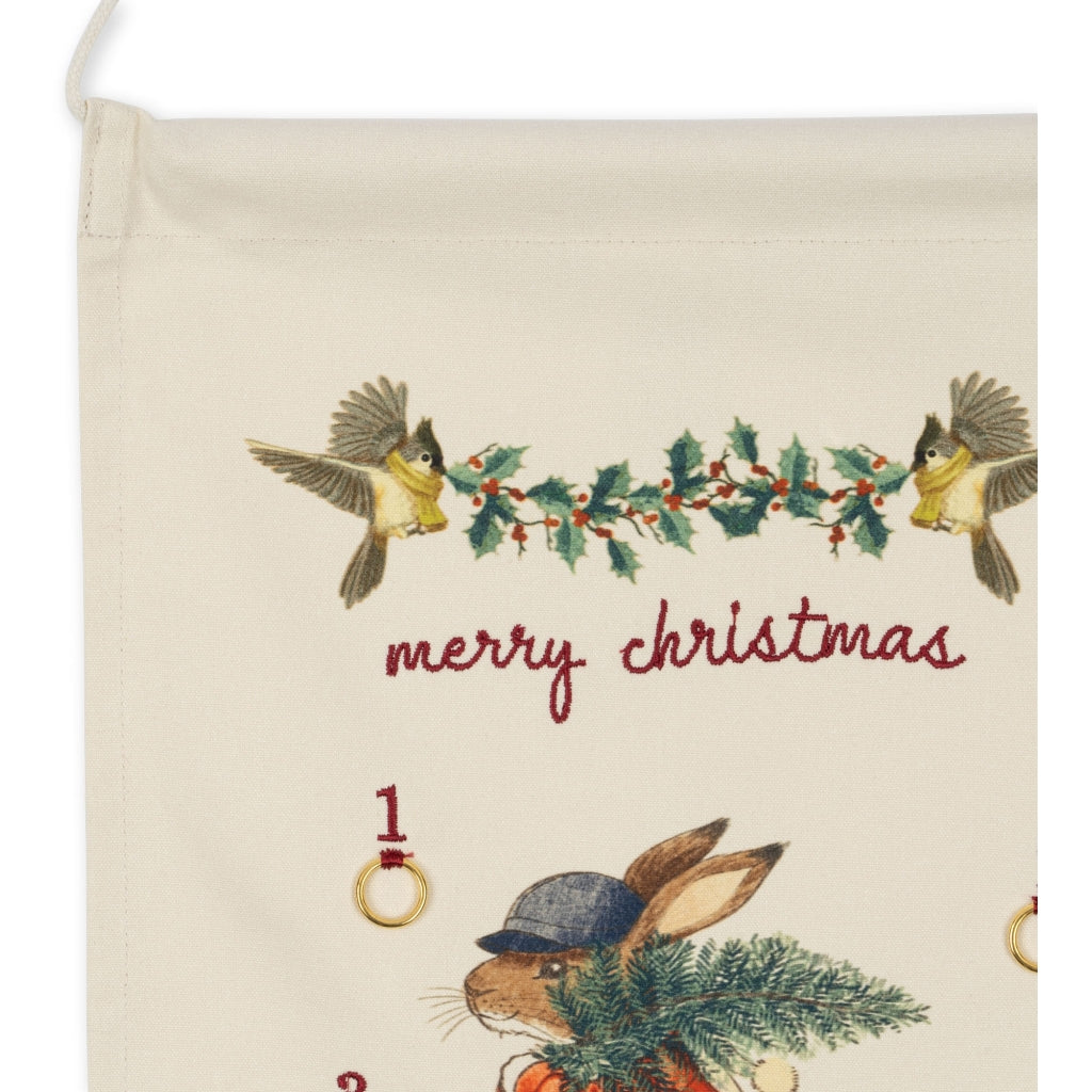 A Christmas Advent Calendar with an embroidered design featuring the text "merry christmas" in red, two birds with holly branches above, and a rabbit holding a pine tree below made from organic cotton.