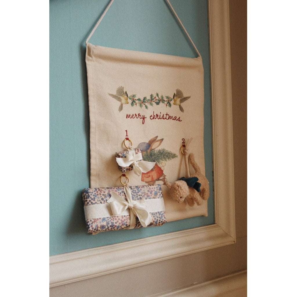 A Christmas Advent Calendar hangs on a wall, featuring pockets decorated with holiday prints and numbers, some filled with small gifts, above the embroidered words "Merry Christmas.