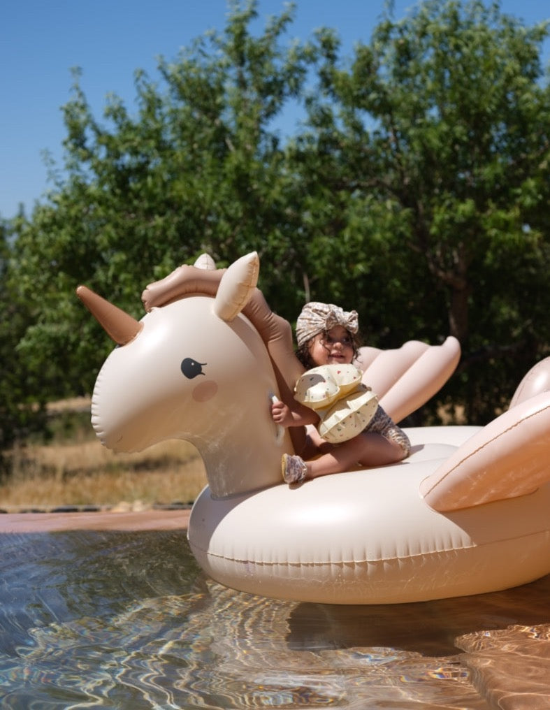 A young child wearing sunglasses and a hat reclines on an Inflatable Unicorn Float in a body of water, surrounded by lush greenery under a clear blue sky.