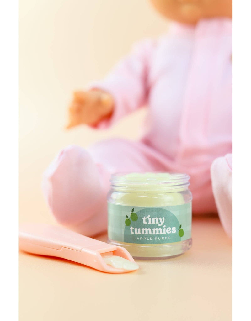 A baby in pink pajamas sits with a jar of "Tiny Tummies - Apple jelly food" and a magic spoon, looking as if a tiny doll is enjoying a meal on the cream-colored background.