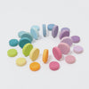 Grimm’s Wooden Pastel Coins, crafted with natural oils, are arranged in a gentle spiral pattern on a white background. The pastel coins display a range of soft shades, including blue, green, yellow, orange, pink, and purple hues.