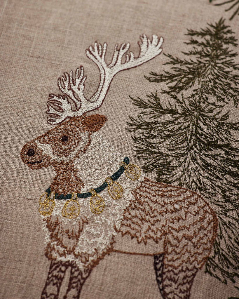 The Coral & Tusk Christmas Cheer Wreath Pillow features an embroidered reindeer wearing a collar with gold bells, standing next to an evergreen tree.