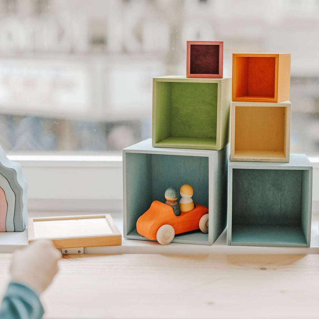 A child's hand reaches towards a set of colorful, non-toxic Grimm's Large Pastel Set of Boxes stacked in a pyramid on a windowsill. One box contains a toy car with two small wooden figures. The background is bright and blurred, highlighting the organized and playful scene featuring this versatile toy perfect for stack and sort fun.