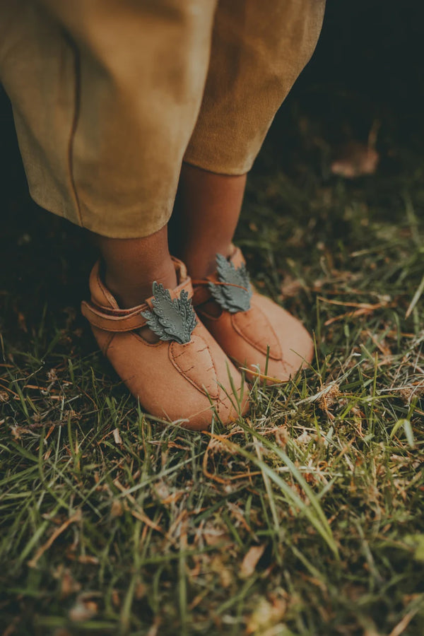 A child wearing tan pants and brown shoes with decorative feather designs stands on soft green grass. The Donsje Baby Carrot Shoes, crafted from premium leather, feature strap closures and are adorned with detailed feather embellishments on the top.