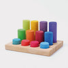 Grimm’s Stacking Game Small Rainbow Rollers consist of ten colorful wooden cylinders of varying heights arranged in a rectangular wooden base with circular cutouts.