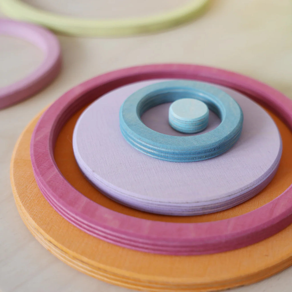 The Grimm's Concentric Circles and Rings Building Set - Pastel features colorful wooden discs and rings in blue, purple, pink, and orange, stacked concentrically for engaging balance games and creative play on a light surface.