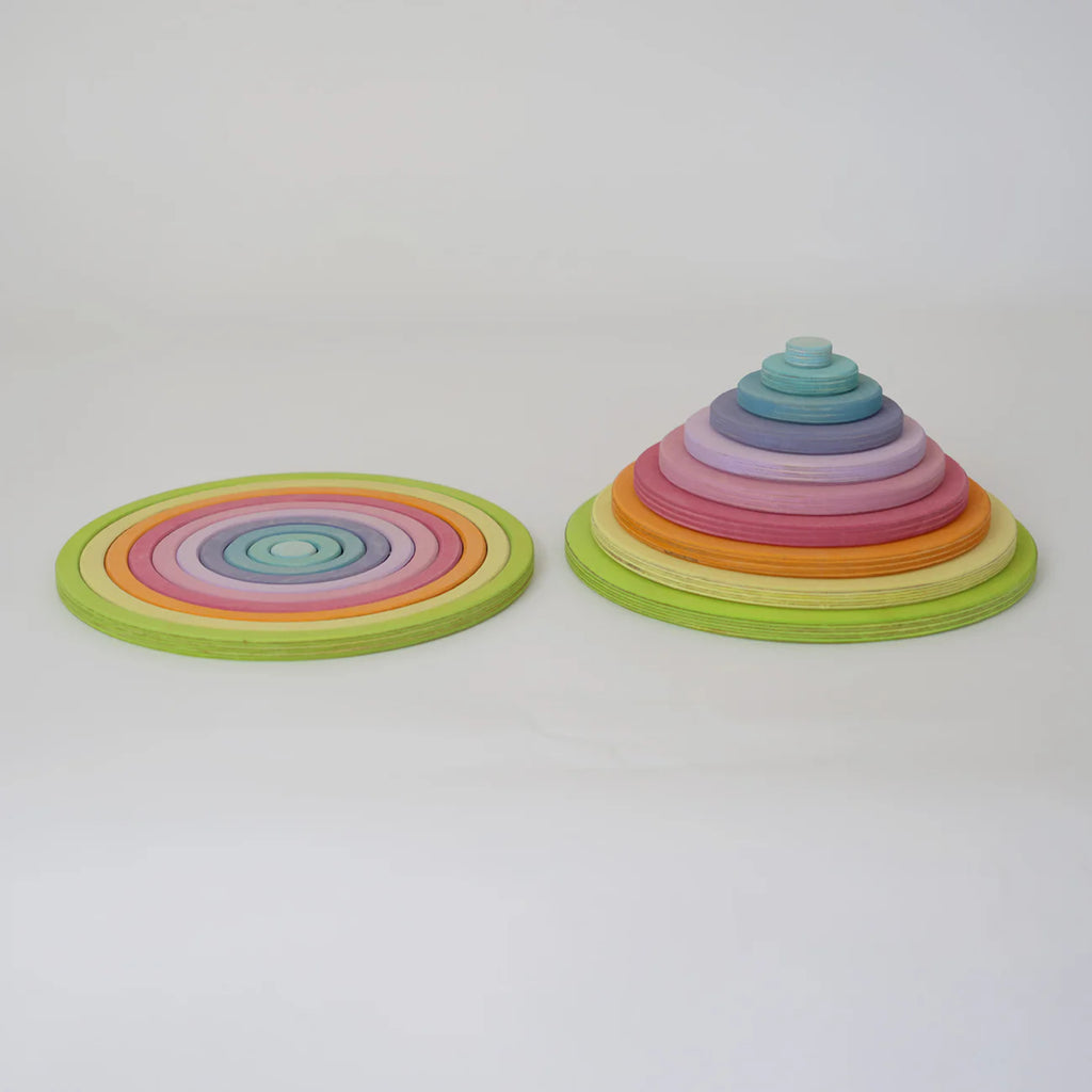 Two sets from Grimm's Concentric Circles and Rings Building Set - Pastel are displayed on a white surface: one arranged in stacked concentric rings, the other built vertically into a cone, showcasing the colorful wooden discs.