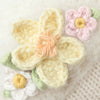 Close-up of crocheted flowers in soft pastels with green leaves on a textured cream background, evoking the delicate handmade details of the Cuddle + Kind Eleanor the Lamb fair trade knit doll from Peru.