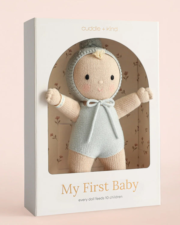 The Cuddle + Kind My First Baby (Bluebell) doll, dressed in a light gray outfit and bonnet, is displayed in a white box with “My First Baby” and “cuddle + kind” on it. The interior of the box features a delicate floral pattern. Ships in one week.