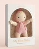 The Cuddle + Kind My First Baby (Rose), a soft hand-knit doll in a pink hood and onesie made from natural cotton yarn, is displayed in a cream box with floral backdrop. Ships in one week.