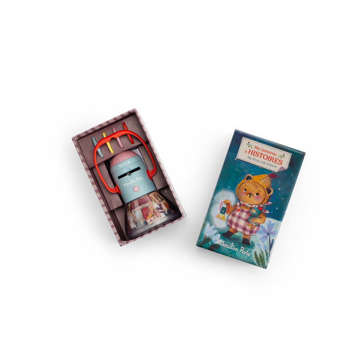 A small toy robot with red headphones sits beside an open box and a closed box featuring a bear holding a lantern, labeled "Moulin Roty Storybook Lantern - The Minouchkas" and "Ma Lampe à Histoires," hinting at magical storytelling.