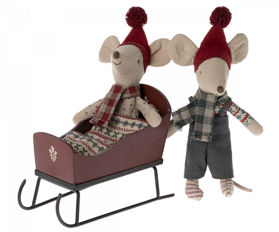 Two plush mice dressed in cozy winter clothes. One mouse is sitting in a Maileg Christmas Sleigh - Red with hand-painted details, wearing a patterned sweater and scarf, while the other stands beside in overalls, striped shirt, scarf, and both wearing red knit hats with pom-poms.