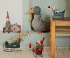 A variety of handmade fabric dolls, including gnomes, a duck, and a mouse, are displayed with dollhouse furniture. Some sit in decorative Maileg Sleighs, Mouse - Green, and a miniature suitcase is next to a gnome on the floor.