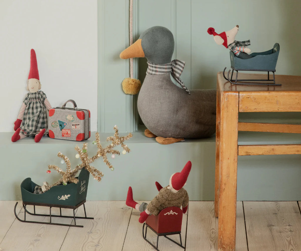 A variety of handmade fabric dolls, including gnomes, a duck, and a mouse, are displayed with dollhouse furniture. Some sit in decorative Maileg Sleighs, Mouse - Green, and a miniature suitcase is next to a gnome on the floor.