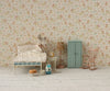 A cute mouse doll sits on a small bed in a cozy, vintage-style room with floral wallpaper, a blue wardrobe, rocking chair, lamp, and tiny breakfast table set. Next to the bed stands the charming Maileg Mini Side Table, adding to the whimsical ambiance of this delightful scene.