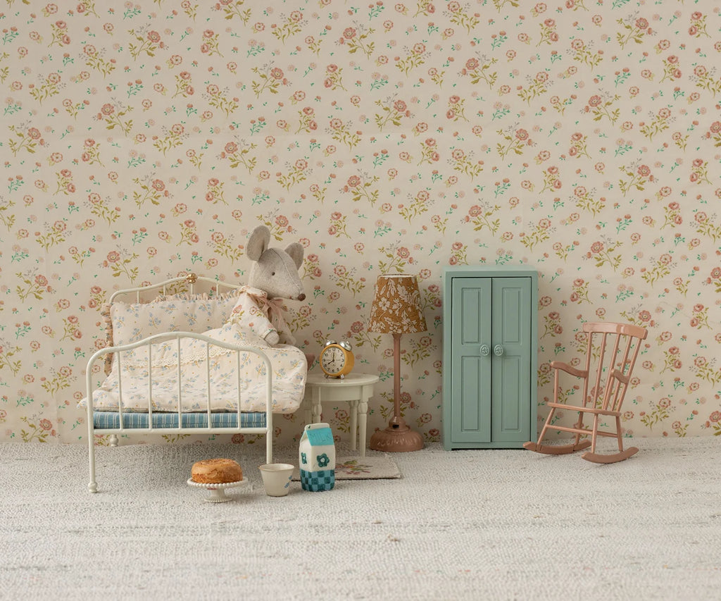A cute mouse doll sits on a small bed in a cozy, vintage-style room with floral wallpaper, a blue wardrobe, rocking chair, lamp, and tiny breakfast table set. Next to the bed stands the charming Maileg Mini Side Table, adding to the whimsical ambiance of this delightful scene.