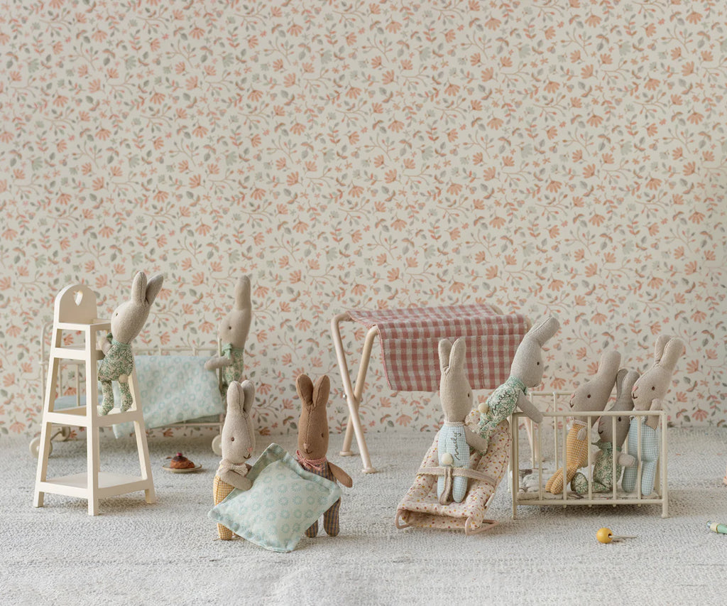 Six small fabric bunny dolls are in a nursery featuring the Maileg Nursery Table, Micro - Red Checker, surrounded by baby furniture, patterned wallpaper, and cozy blankets for a playful, charming scene.