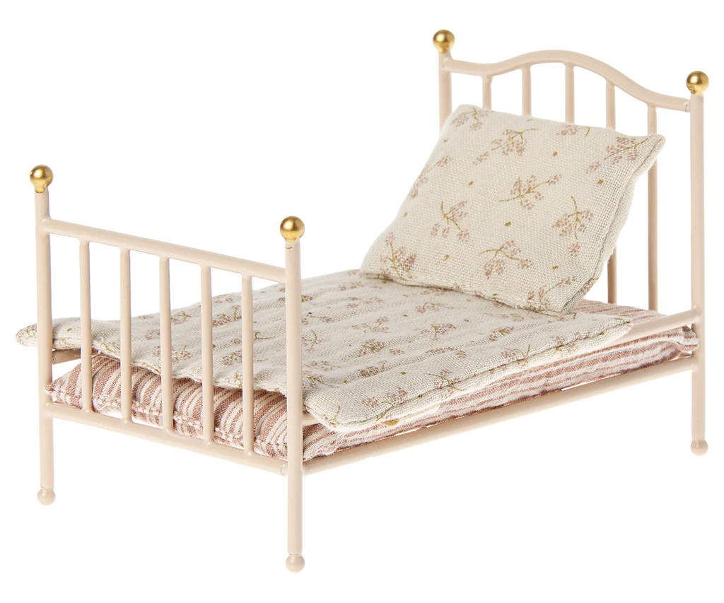 The Maileg Little Sister Bedroom Starter Set includes a small, vintage-style metal bed frame with gold accents, perfect for collectors. It comes with a floral-patterned pillow and mattress. The light-colored bedding features a subtle design, creating a quaint and cozy look.