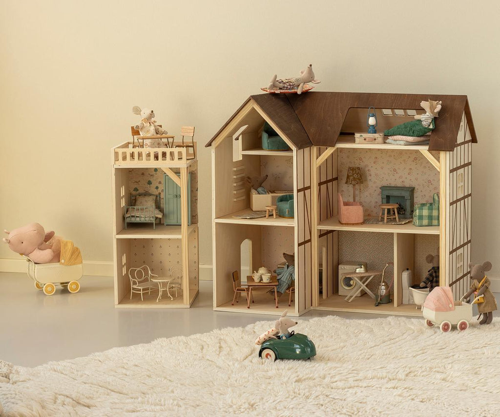 Two wooden dollhouses, including the charming Maileg Farmhouse Dollhouse, with miniature furniture and toy mice are arranged on a white rug. Nearby, a small toy car and a toy pig in a stroller add to the cozy, playful atmosphere, evoking a delightful mouse dollhouse world.