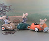 Three Maileg Mouse Car - Light Brown, each dressed in cute outfits, riding in colorful miniature metal cars on a tabletop, with a soft focus background simulating a landscape.