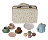 A set from the Maileg collection displayed: on the left, plaid shorts featuring brown, black, and white stripes; in the center, a beige and brown diamond-patterned tie; on the right, a cozy brown cardigan. Behind these clothes is a charming small suitcase adorned with hotel stickers. Perfect for sparking creativity and storytelling!