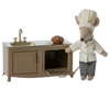A Maileg Kitchen - Light Brown mouse dressed as a chef standing next to a miniature metal kitchen counter with a sink, holding a small baked good. The cabinet door below the sink is open, revealing a tray.