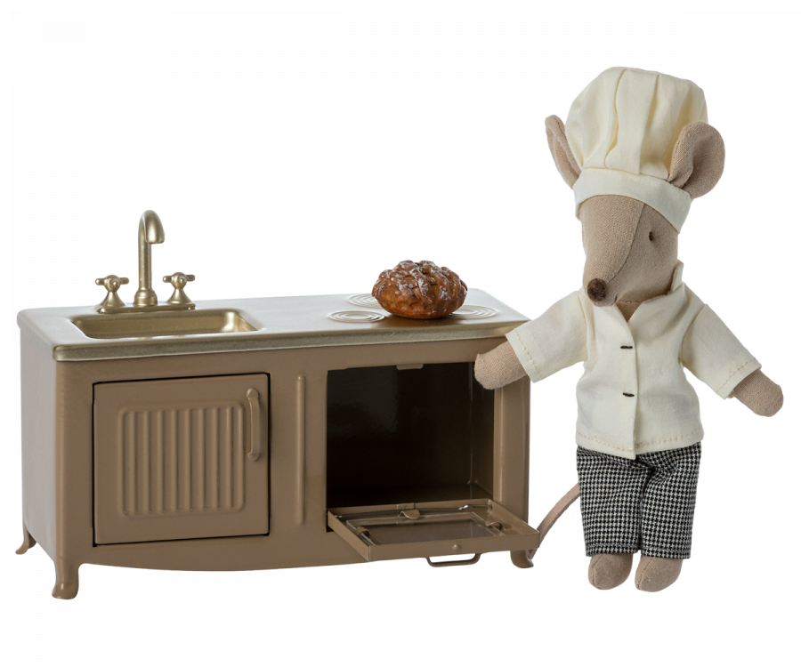 A Maileg Kitchen - Light Brown mouse dressed as a chef standing next to a miniature metal kitchen counter with a sink, holding a small baked good. The cabinet door below the sink is open, revealing a tray.