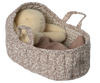 A plush teddy bear is cozy in a large, off-white Maileg Carry Cot with two handles, resembling a portable bed. A warm knitted blanket rests beside the bear.