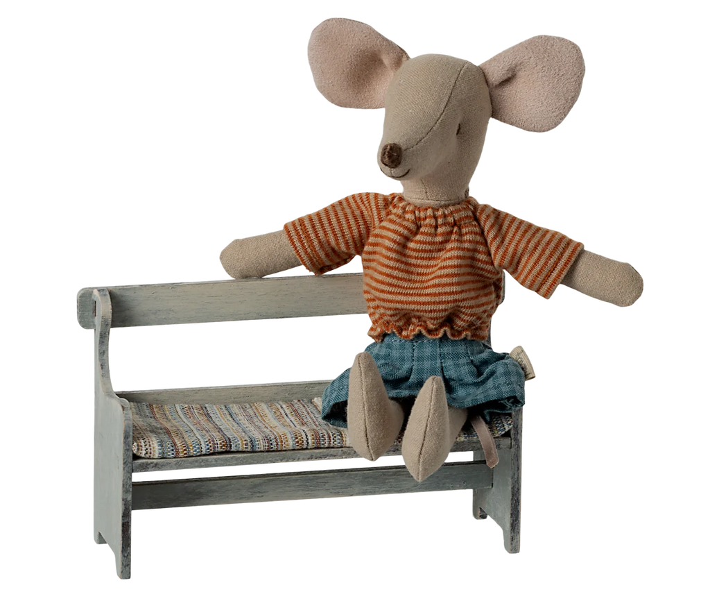 An elegantly poised plush mouse toy dressed in a striped orange shirt and denim skirt rests on the Maileg Mouse Bench in off-white, which features an intricately hand-painted design. A cushion with vibrant, colorful stripes provides added comfort to the scene.