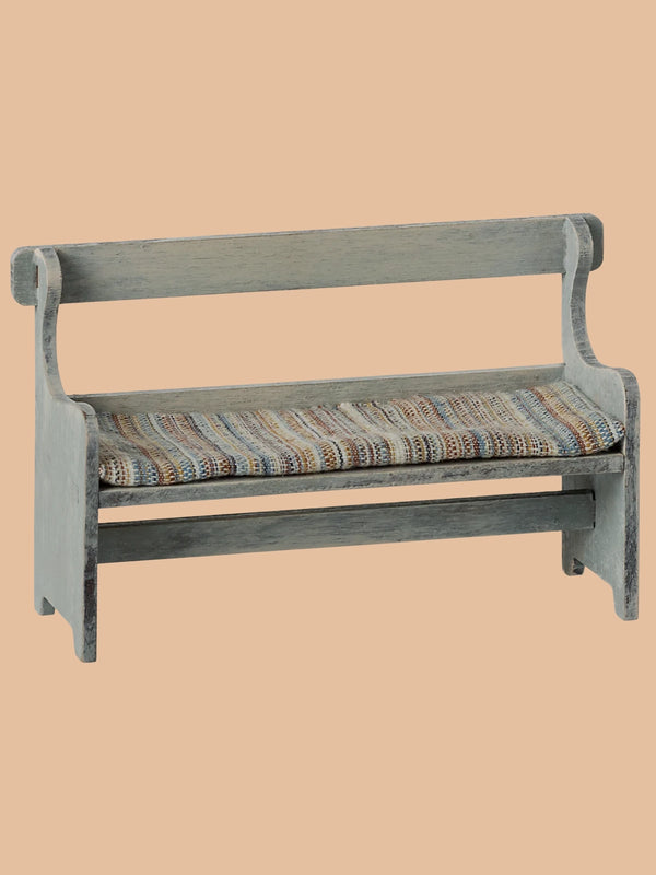 The Maileg Mouse Bench - Off-White is a rustic wooden bench with hand-painted details. It includes a backrest and a woven, multicolored fabric seat cushion, accentuated by charming curved armrests against a plain beige background.