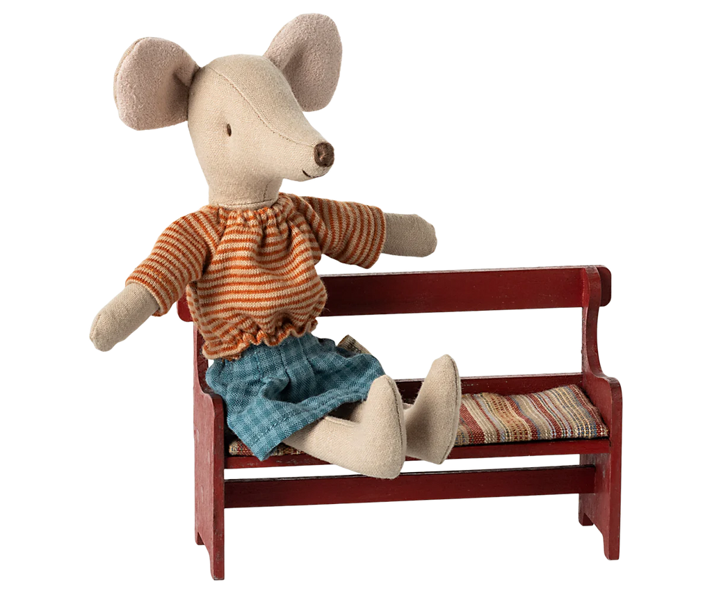 A charming plush mouse with a vintage flair is outfitted in an orange striped shirt and blue shorts. It relaxes on the Maileg Mouse Bench - Red, which includes a patterned seat cushion, with one arm draped casually over the back. Its large ears hint that it belongs to an adorable mouse family.