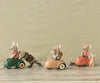 Three Maileg mouse cars, each uniquely clothed, are engaged in mouse transportation in small, colorful vintage metal cars against a patterned background. One mouse pulls a smaller mouse in a pull toy cart.