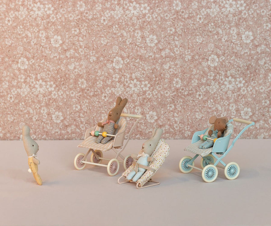 Three Maileg Stroller, Baby Mice - Rose, each in a different colored vintage stroller, are positioned on a surface against a floral background. Their outfits and strollers vary in color from pink to blue.