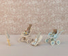 Three rabbit dolls, each in a Maileg Stroller, Baby Mice - Blue, are positioned on a floral patterned background. The strollers are light blue, beige, and blue with polka dot fabric. A fourth smaller rabbit doll walks towards them carrying a toy carrot, reminiscent of how mouse families transport babies.