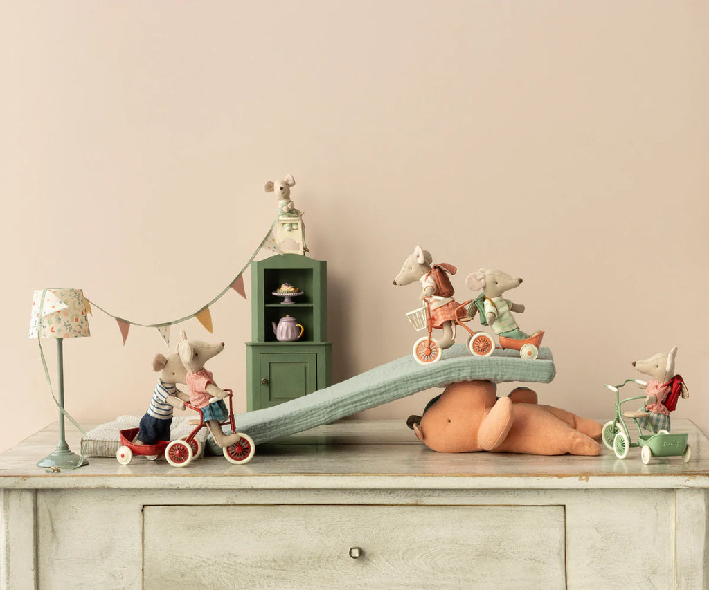 A whimsical scene featuring toy mice on vintage-style tricycles with tiny, intricate tricycle handles riding down a small ramp on a table. Maileg Big Sister With Backpack - Red stands nearby, observing. A teddy bear lies beneath the ramp. The background has a small green cabinet with miniature tea sets and decorations, evoking the charm of kindergarten days.