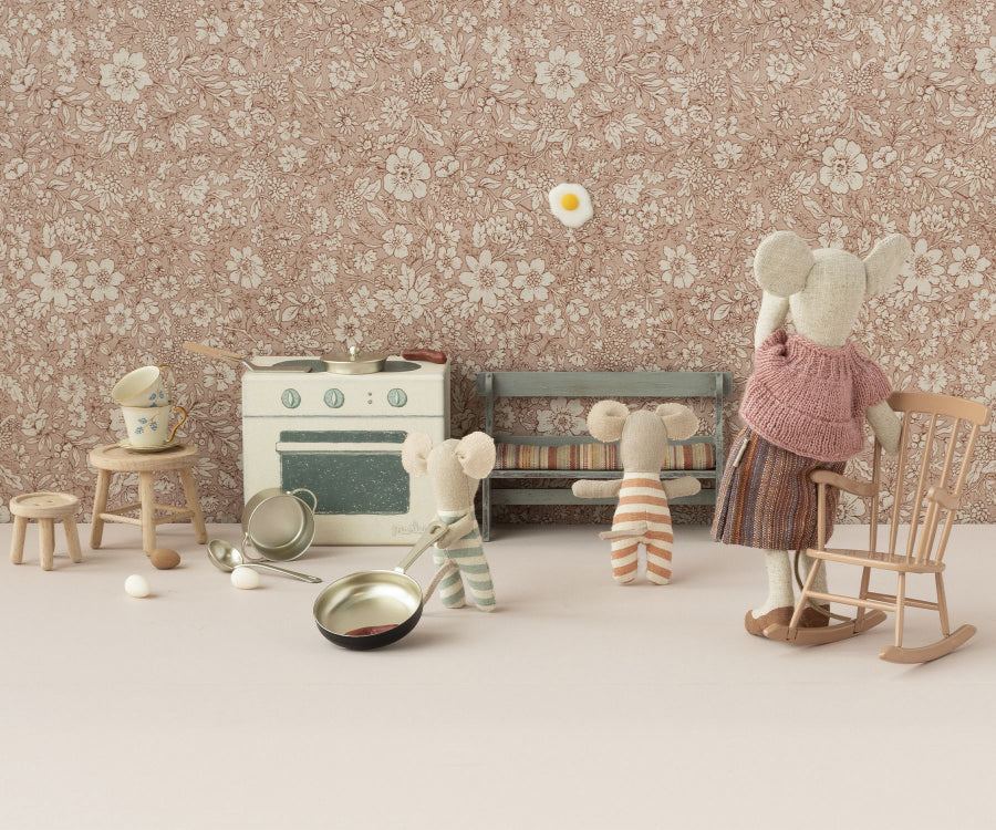 A whimsical scene with three stuffed mice in a miniature kitchen. One mouse in a pink sweater flips an egg in a frying pan beside a Maileg Rocking Chair, Mini - Dark Powder (Larger Size). Two smaller mice are nearby, one reaching up, the other holding a wooden spoon. The kitchen has a stove, utensils, and floral wallpaper.