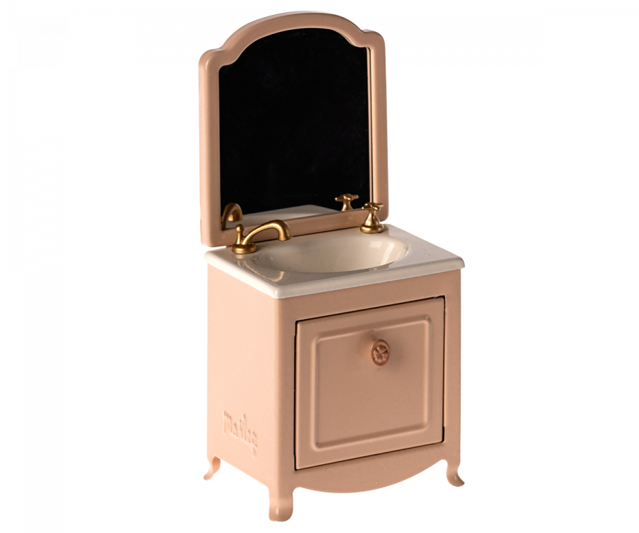 A small model of a vanity sink with a mirror. The Maileg Sink Dresser With Mirror, Mouse features a white countertop, gold faucet, and cabinet door. The mirror, framed in pink, is removable for easy cleaning. Perfect for adding a touch of elegance to your Maileg mouse castle's royal bathroom with its vintage design.