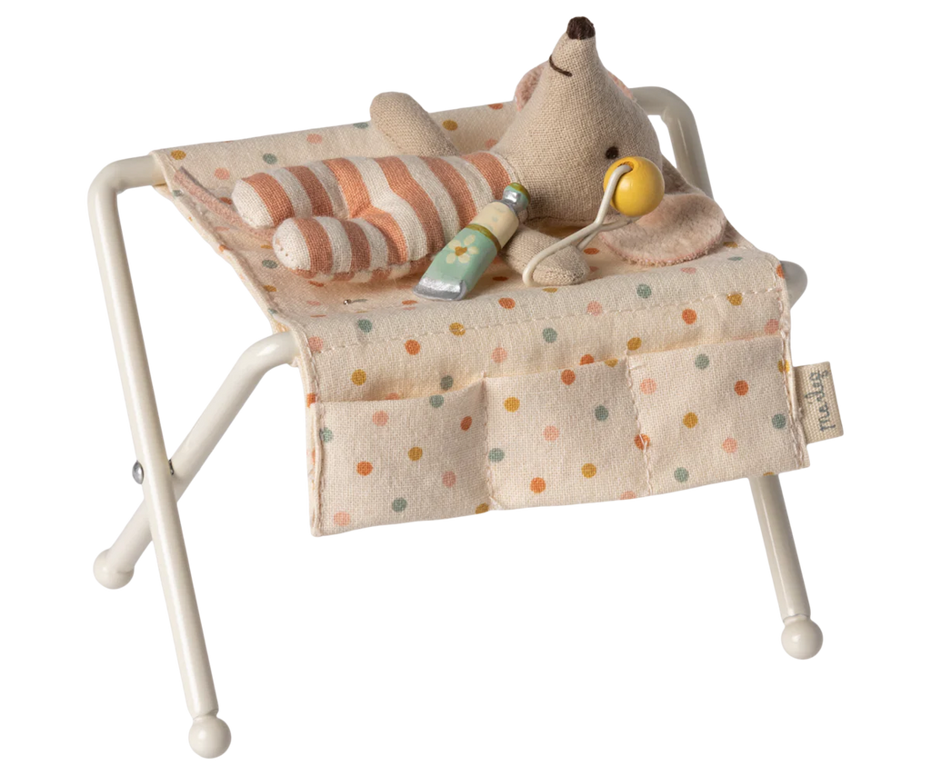 A small stuffed toy mouse lies on the Maileg Nursery Table, Baby Mouse - Off White, adorned in a striped pink and white onesie. Beside it, there is a green baby bottle and a yellow pacifier. The table cover features multi-colored polka dots, evoking the playful charm of a nursery table for baby mice.