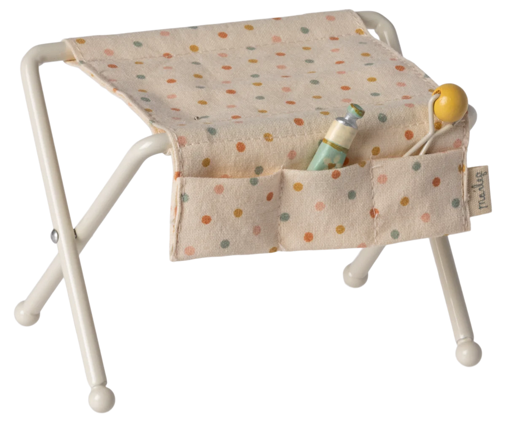 A small white baby bassinet with a polka dot patterned quilt. The quilt has small pockets on its side, one containing a baby bottle and the other containing a baby rattle. Nearby, the Maileg Nursery Table in off white for a baby mouse is thoughtfully arranged with essentials for easy access. The bassinet legs are crossed for support.