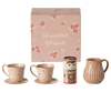 The Maileg Hot Chocolate Set, Mini comes in a beautiful pink gift box adorned with a floral design and the words "Chocolat Chaud." Included in this charming set are two pink mugs with gold polka dots and rims, complete with matching saucers, a brown milk pitcher, and a canister of hot chocolate mix. It's the perfect addition for sharing sweet moments with the Teddy family.